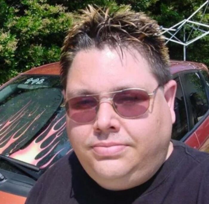 Man with spiked bleached hair and tinted sunglasses standing by a car with flame decals, showing cringey nineties fashion style.