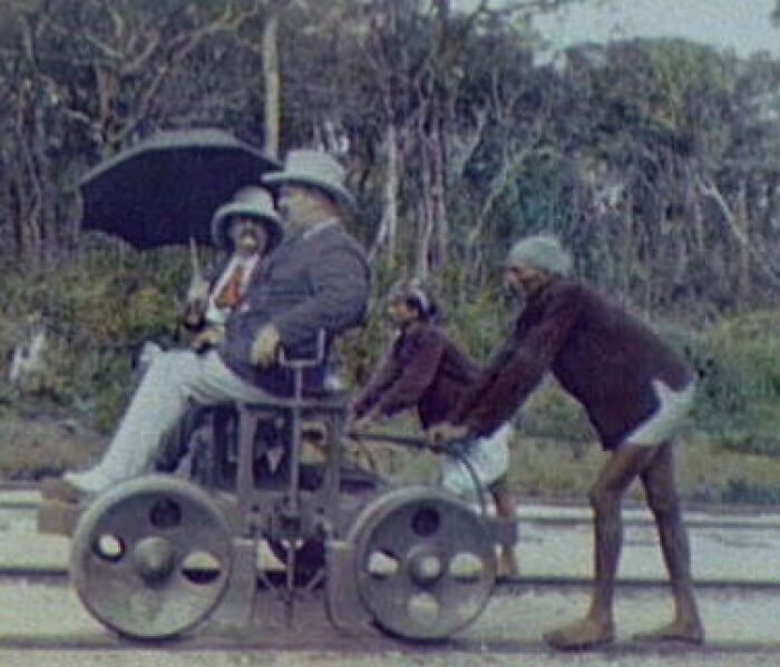Old-fashioned manual railway cart with two workers pushing and two passengers riding in a historic commute scene.