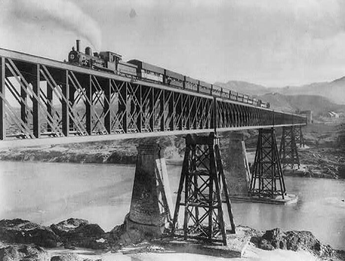 Historic commutes shown by a steam train crossing a tall iron bridge over a river in a bygone working world.