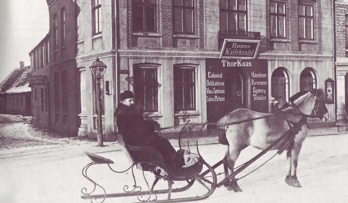 Historic commute scene with a man in a horse-drawn sleigh passing a brick building on a snowy street.