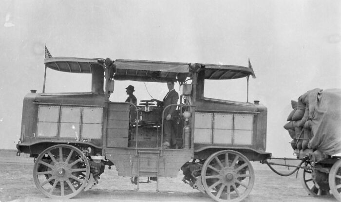 Early 20th-century historic commute showing men riding in a vintage motorized vehicle pulling a loaded cart.