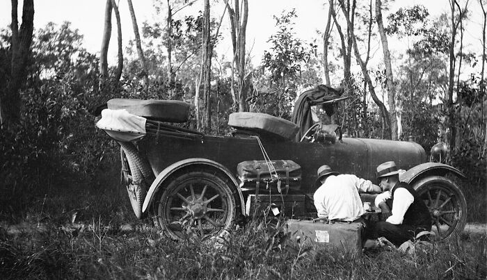 Two men repairing an early automobile on the roadside, illustrating historic commutes in a bygone working world.