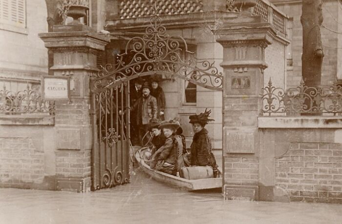 Historic commutes showing people in a small boat navigating flooded streets near an ornate wrought iron gate.