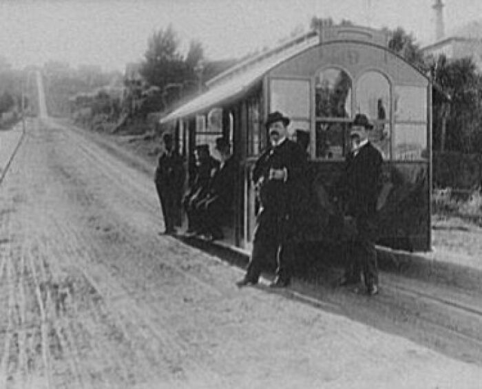 Historic commutes with early 20th-century workers standing by a vintage railcar on a rural dirt road.