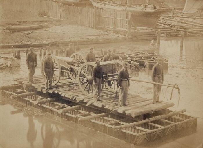 Men on a wooden raft transporting a cannon and cart over water in a historic commute from a bygone working world.