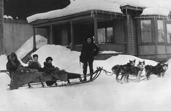 Historic commutes showing a dog sled with passengers and driver in deep snow outside a wooden building.
