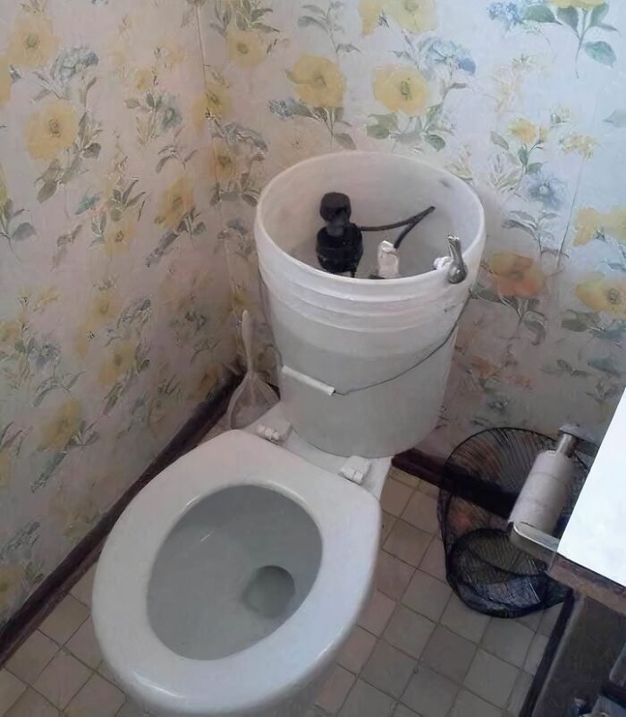 Toilet with a bucket used as a tank in a bathroom with floral wallpaper, an example of worst real estate photos ever.