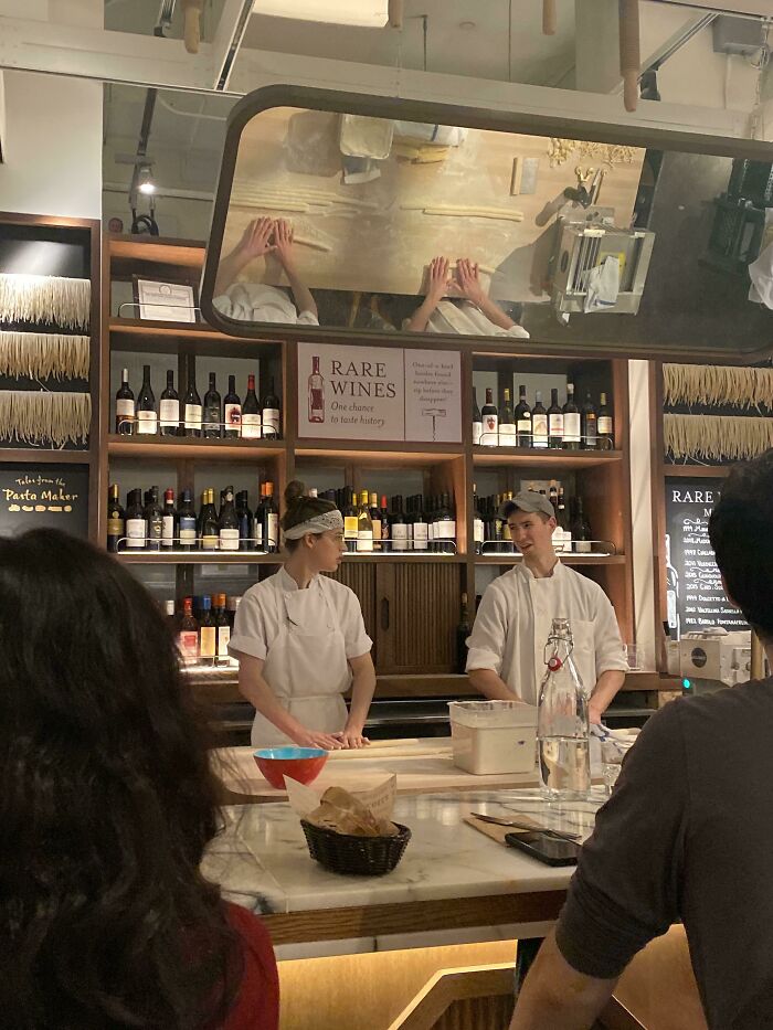 Two chefs demonstrating clever pasta-making techniques in a kitchen with wine bottles on shelves behind them.