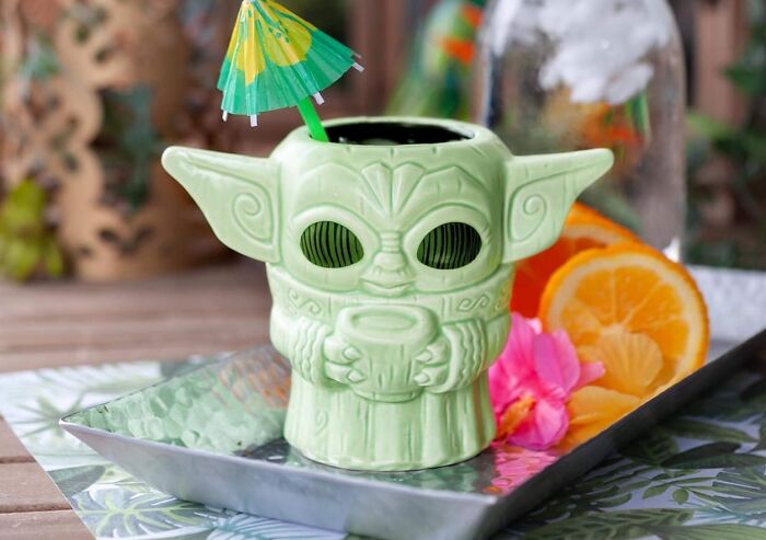 Green ceramic mug shaped like a character holding a cup, with a tropical umbrella and orange slices on a silver tray.