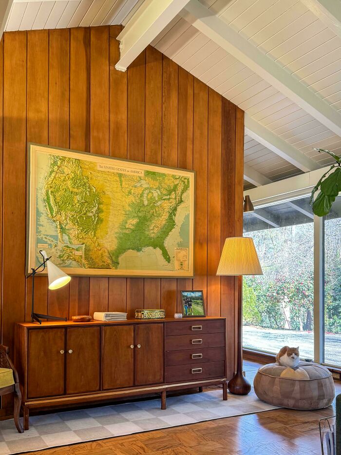 Mid-century vintage living room with wooden sideboard, map wall art, floor lamp, and cat on pouf by large window.