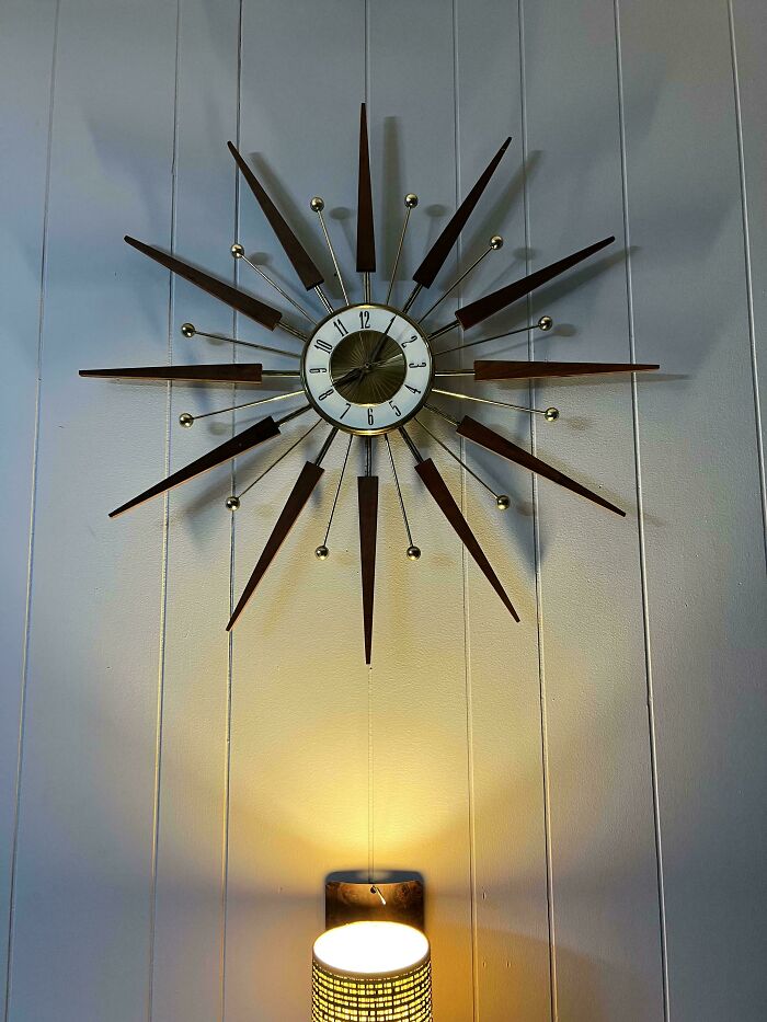 Mid-century design starburst wall clock above a lit vintage lamp on a white paneled wall.