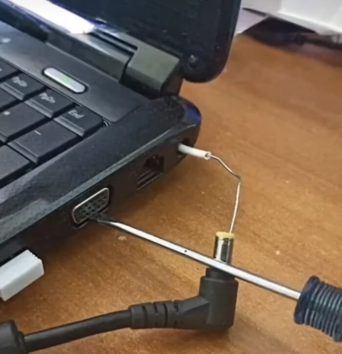 Laptop with a makeshift technology disaster repair using a screwdriver and a bent wire on a wooden surface.