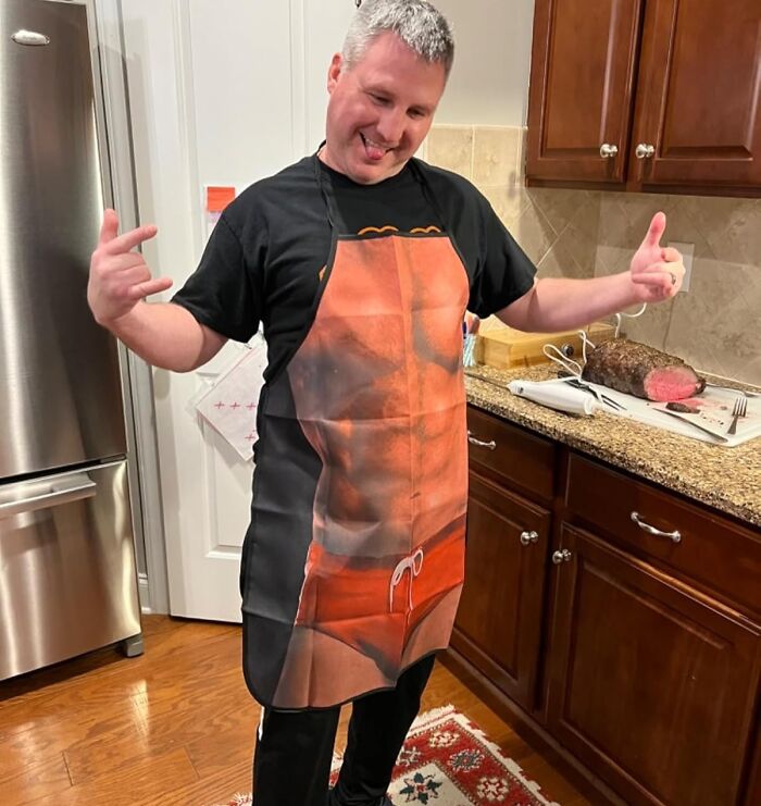 Man wearing a funny kitchen gadget apron with a printed muscular torso, standing in a kitchen with a roast on the counter.