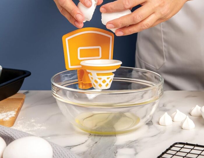Mini basketball hoop kitchen gadget used to crack eggs into a mixing bowl on a marble countertop.