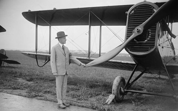 Man in vintage suit and hat standing next to propeller of an old biplane in a classic aviation photo.