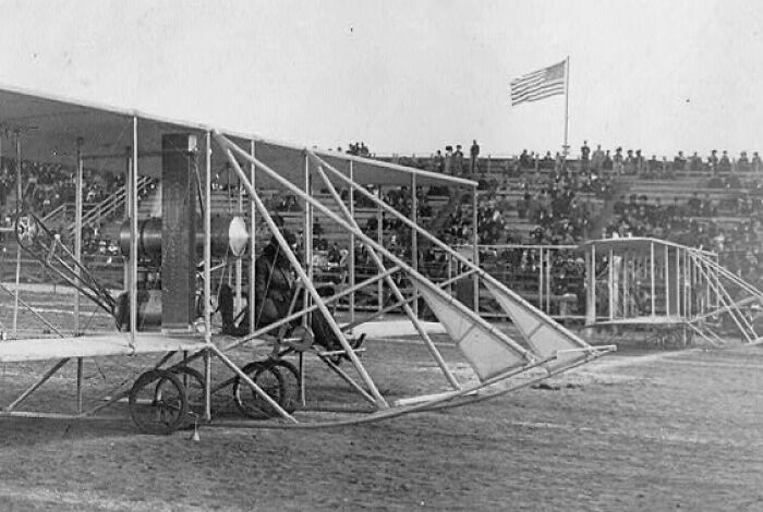 Vintage photo of an early plane on a dirt runway with spectators in stands, capturing the magic of aviation history.