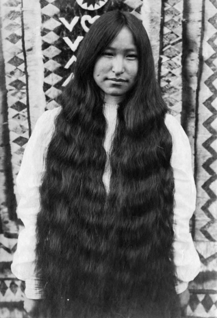 Black and white photograph of Native American female with long wavy hair standing in front of patterned textile, honoring strength.