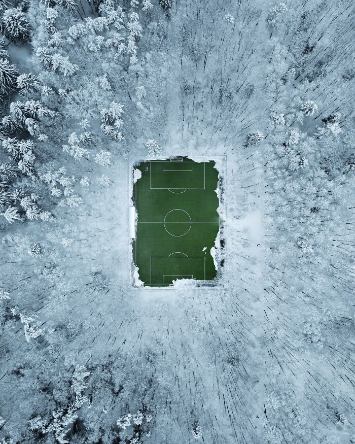 Aerial photo of a snowy forest surrounding a green soccer field, showcasing stunning aerial photography by Borsch.