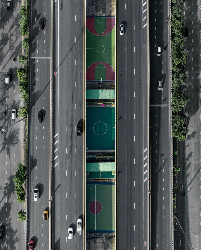 Aerial photo showing multiple colorful sports courts between two busy highways with cars and green trees lining the roads.