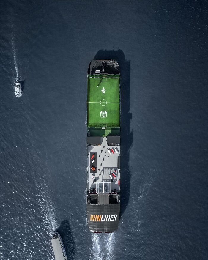 Aerial photo of a ship with a soccer field on deck sailing through deep blue water surrounded by two smaller boats.