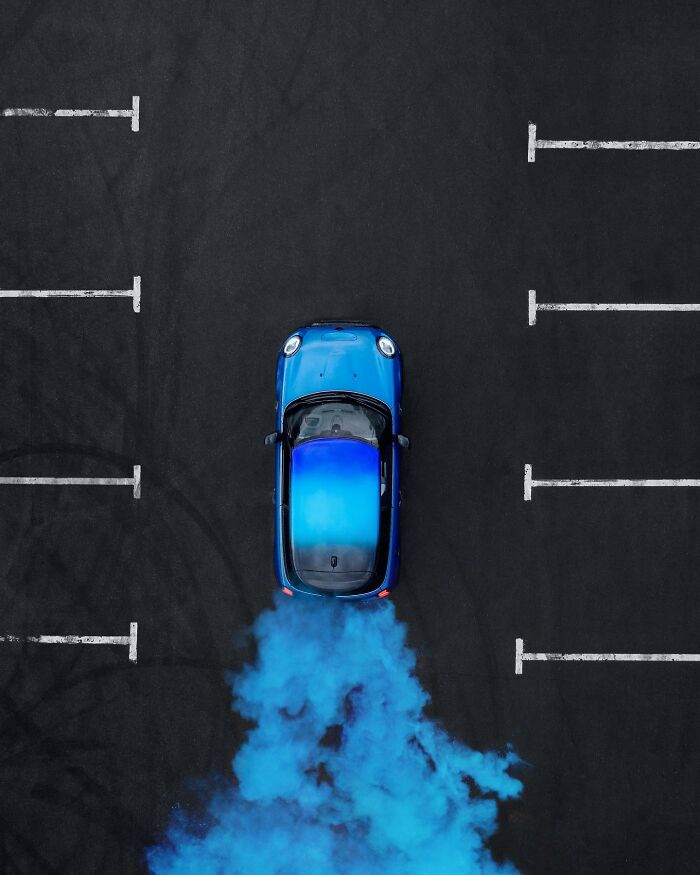 Aerial view of a blue car emitting blue smoke in an empty parking lot from above, unique aerial photo perspective.
