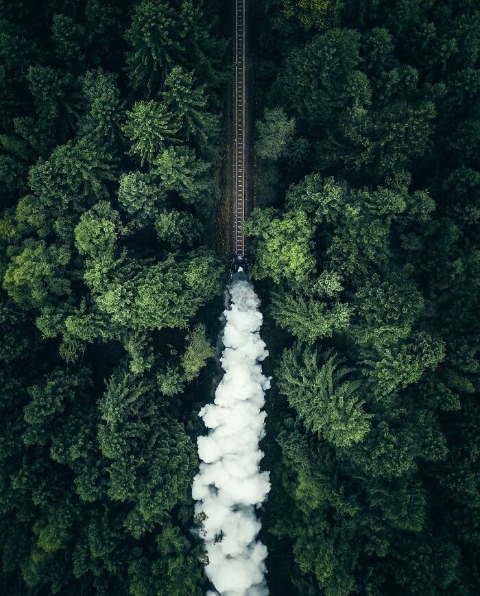 Aerial view of a train moving through dense green forest leaving a trail of white smoke in aerial photos.
