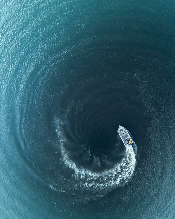 Aerial photo showing a small boat creating a circular wake pattern on deep blue water from above.