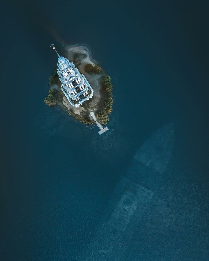 Aerial photo of a white tower on a small island with surrounding deep blue water and a sunken ship visible below.