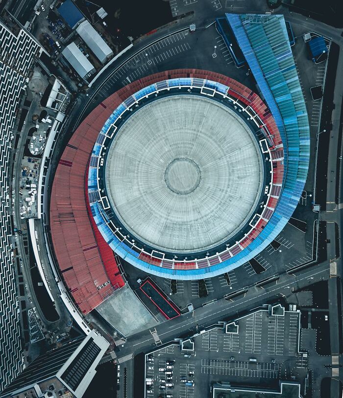 Aerial photo of a circular stadium with red and blue seating, surrounded by parking lots and urban structures.