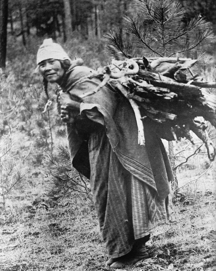 Native American woman carrying firewood in a time-weathered photograph honoring female strength in a forest setting.