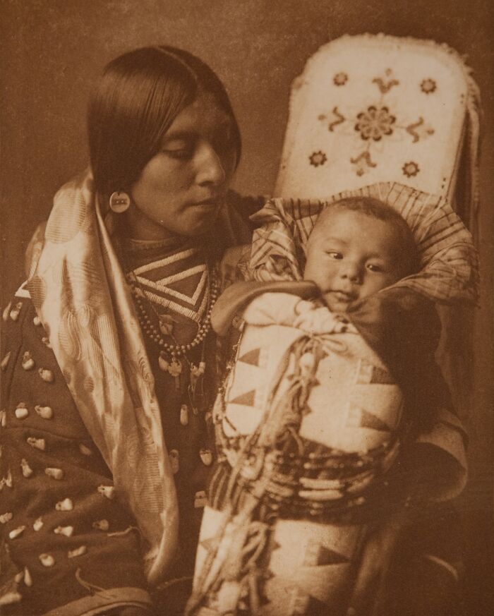 Native American woman holding baby wrapped in traditional clothing, showcasing female strength in a time-weathered photograph.
