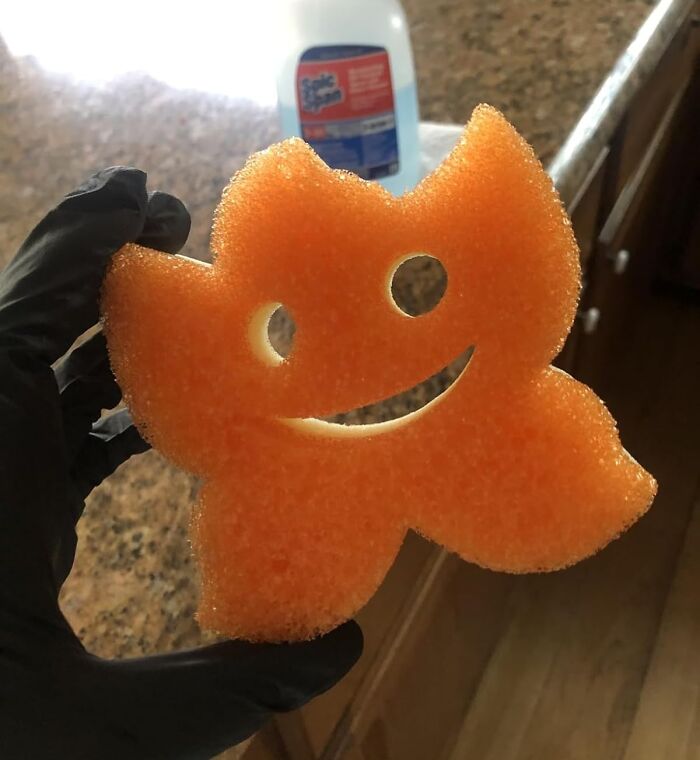 Orange smiling kitchen sponge held by hand wearing black glove, popular Shark Tank product for cleaning and scrubbing tasks.