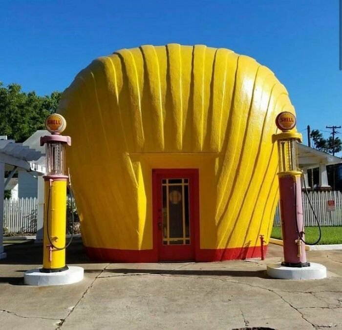 Vintage yellow Shell gas station shaped like a large seashell with two old-fashioned gas pumps under clear blue sky.