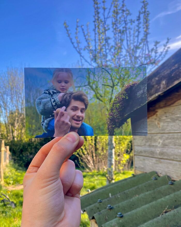 Hand holding a movie scene photo aligned with the real-life filming location in a garden setting during daytime.