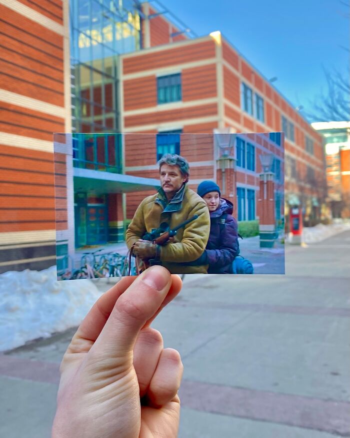 Hand holding a photo of movie scene with two characters matched to the real-life filming location outside a brick building.