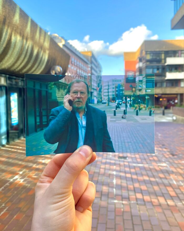 Hand holding a photo showing a real-life filming location of a movie scene in an urban city setting with buildings and pedestrians.
