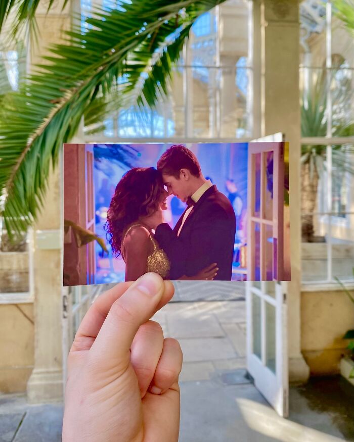 Hand holding a photo of a movie scene at a real-life filming location with plants and glass windows in the background.