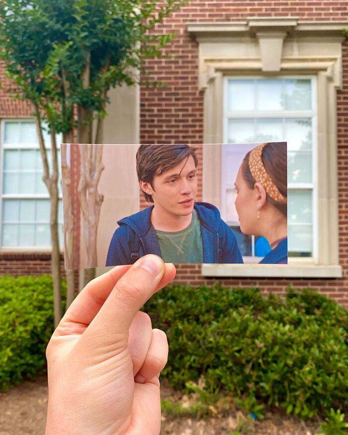 Hand holding photo of movie scene in front of real-life filming location with brick building and greenery behind.