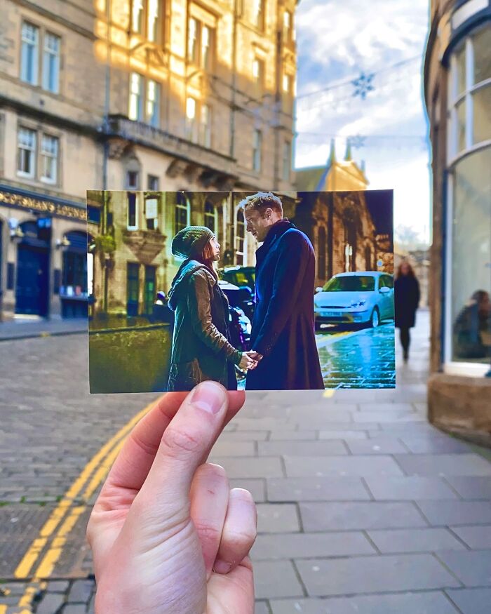 Hand holding a movie scene photo matching the real-life filming location on a city street.