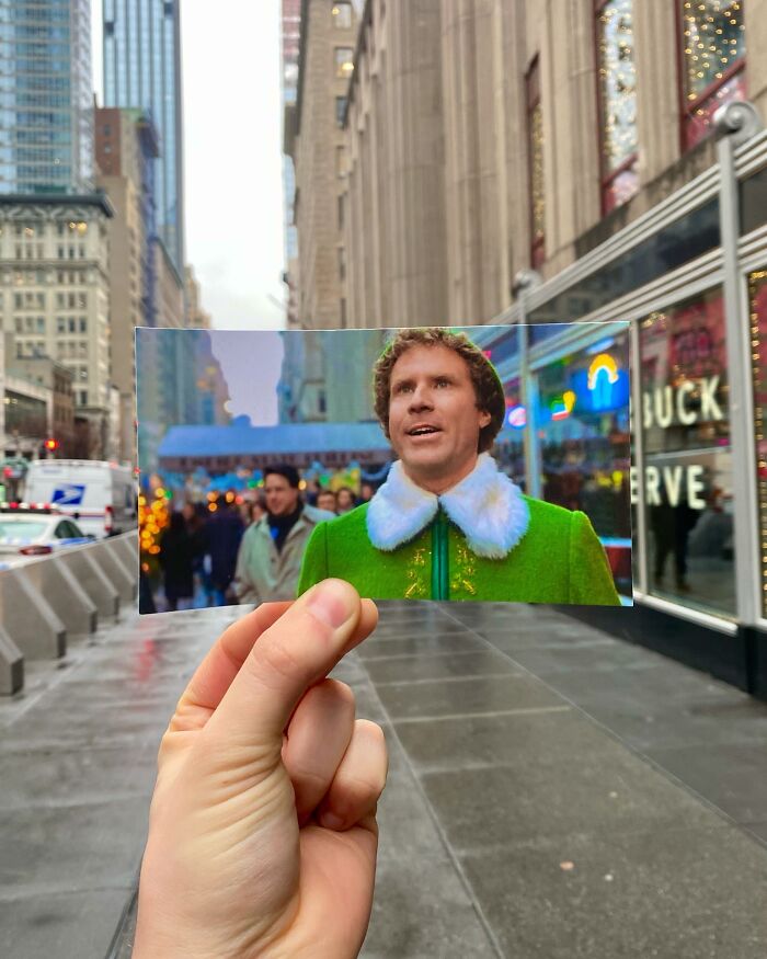 Hand holding photo of Elf movie scene in real-life filming location on city street with pedestrians and buildings.