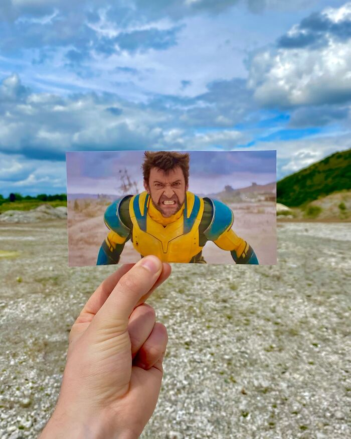 Hand holding movie scene photo in front of the real-life filming location of a superhero in colorful costume outdoors.