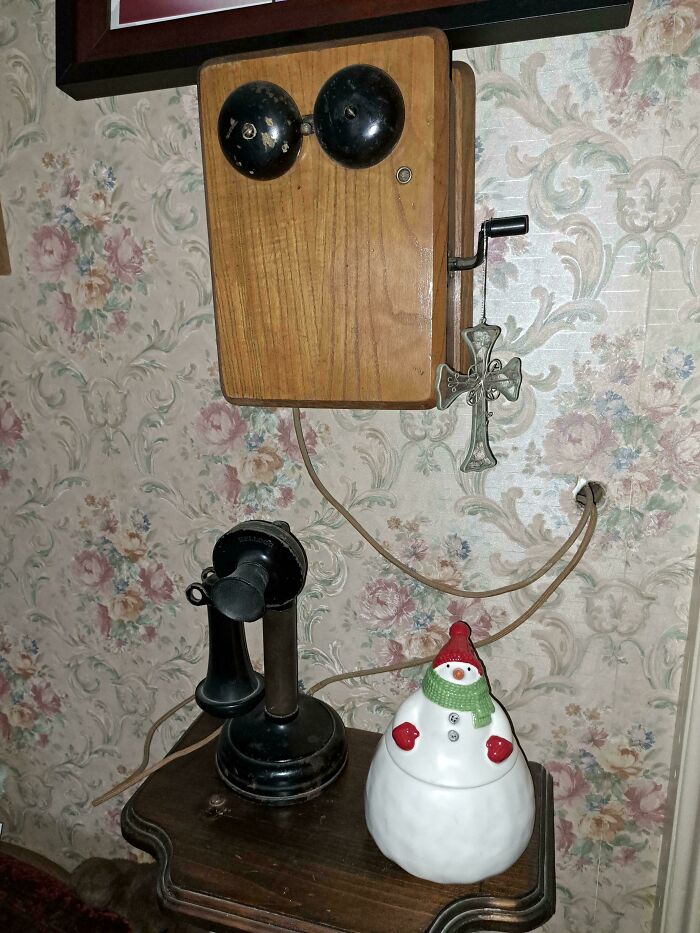 Vintage wooden wall telephone with a crank and black candlestick handset on a wooden table with floral wallpaper background