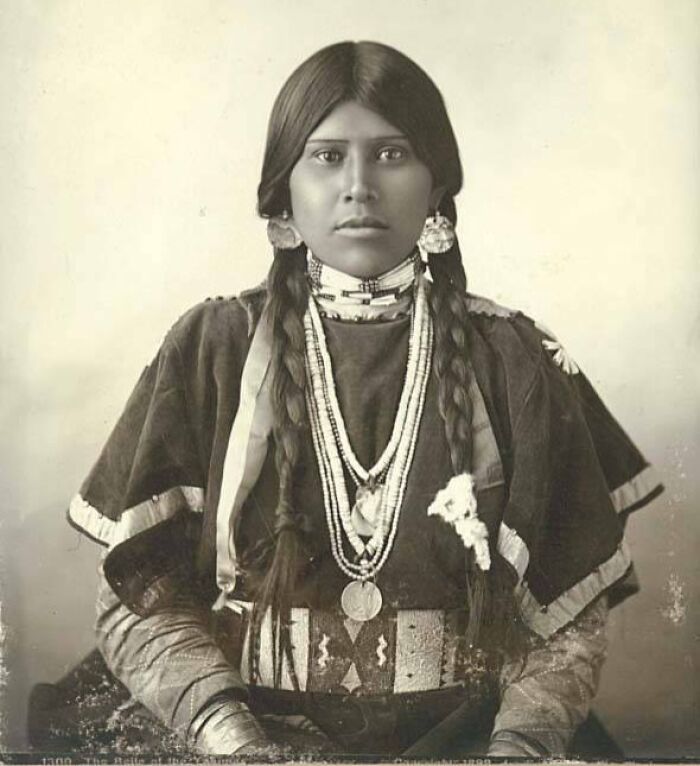 Native American woman in traditional attire with braids and jewelry, showcasing female strength in a time-weathered photograph