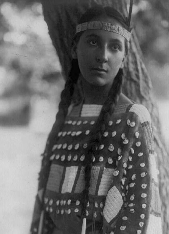 Native American woman in traditional attire with long braids standing outdoors near a tree honoring female strength.