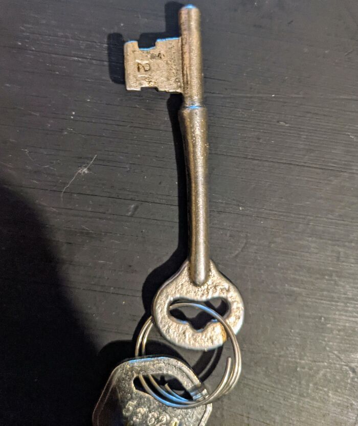 Old rusty metal key with unique shape and attached modern keys on a dark wooden surface in an old house.