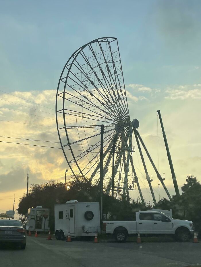 Ferris wheel under construction at sunset with vehicles parked nearby in a scene for curious minds.