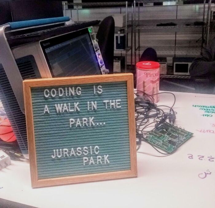Letter board on office desk with funny coding quote, surrounded by electronics and a red container, showcasing office personality finds.