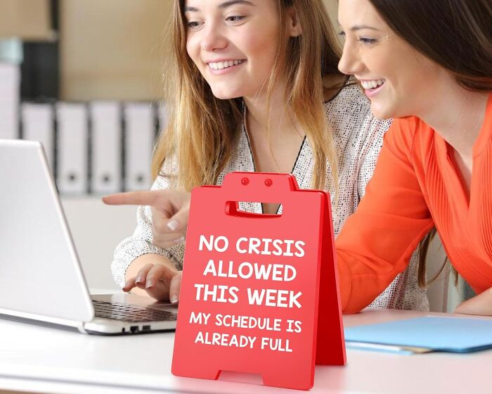 Two women smiling at a laptop with a red funny office sign about no crisis on a busy schedule nearby.