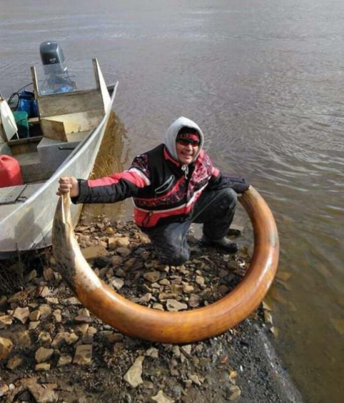 Man holding a large historic ivory tusk by a riverbank next to a fishing boat, a bizarre historical artifact display.