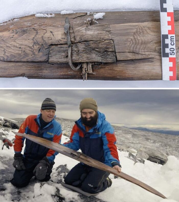 Two men outdoors in snow holding a large bizarre historical artifact made of wood with metal parts visible.